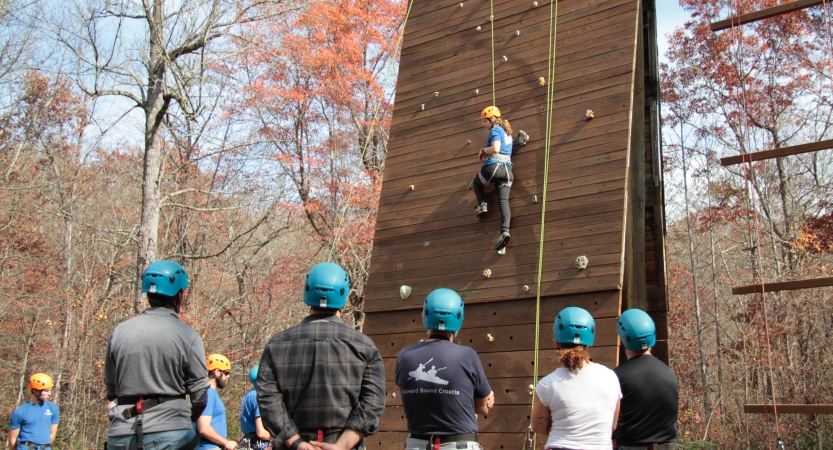 A person wearing safety gear is secured by ropes while participating in a ropes course. Others wearing helmets below look on. 